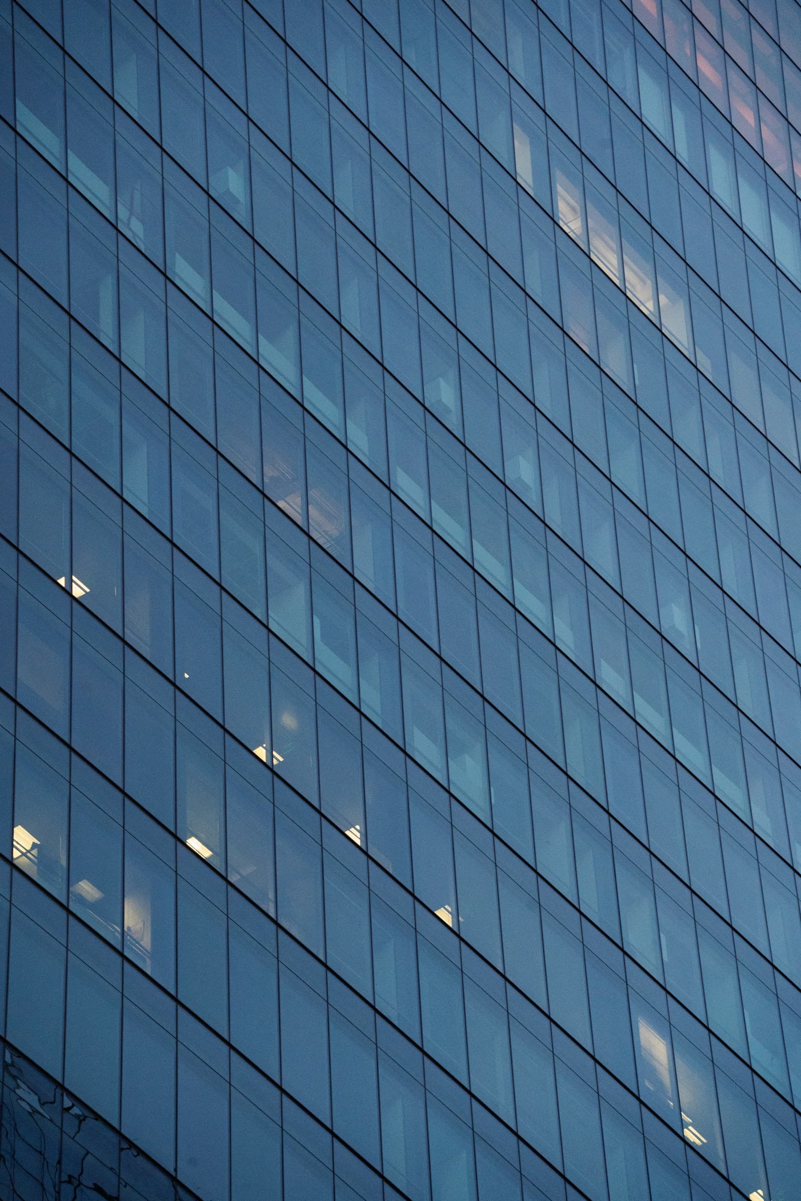The glass facade of a modern office building, its panels catching scattered interior lights